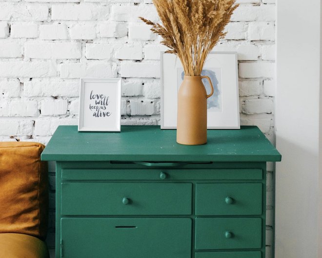 A dark green dresser with framed photos and a vase on top, set against a white brick wall.