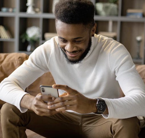 A man is sitting on a light brown sofa, smiling and texting on his phone. There are black shelves with various items in the background.