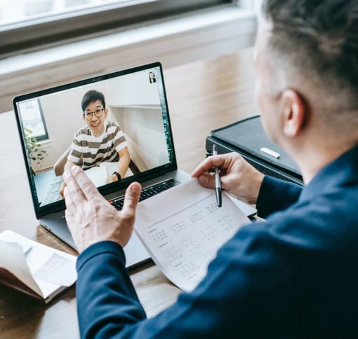 A computer on a wooden desk displays a video call of a smiling man with a stack of papers in front of him. In the foreground, a man is seen taking notes while engaged in the video call.