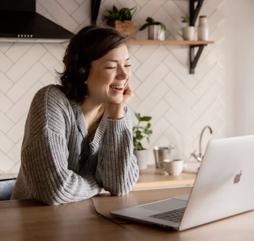 A woman wearing a gray sweater is smiling during a video call on a MacBook. She is seated at a wooden desk, with a white wall and wooden shelves in the background.