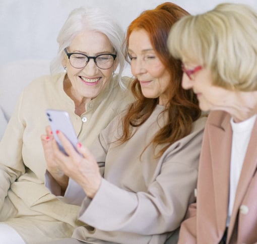 Three elderly women in light cream attire happily engaged in a video call on a mobile phone, held by the woman in the middle.