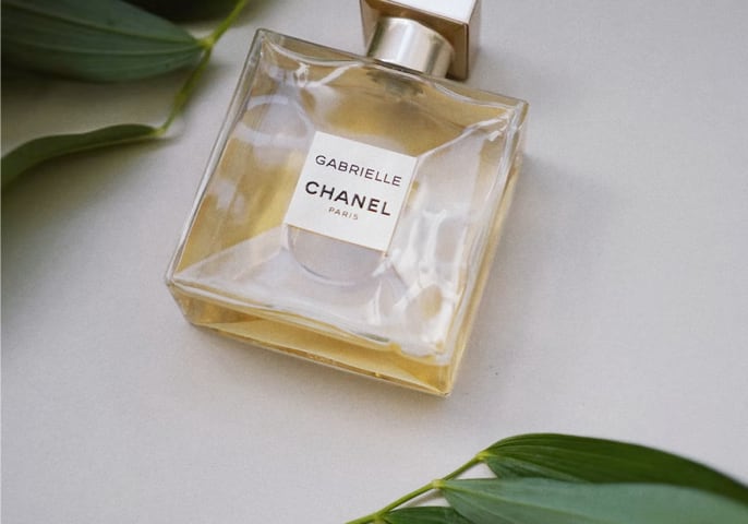 A bottle of Gabrielle Essence Eau De Parfum lays on a white surface between two leafy stems.